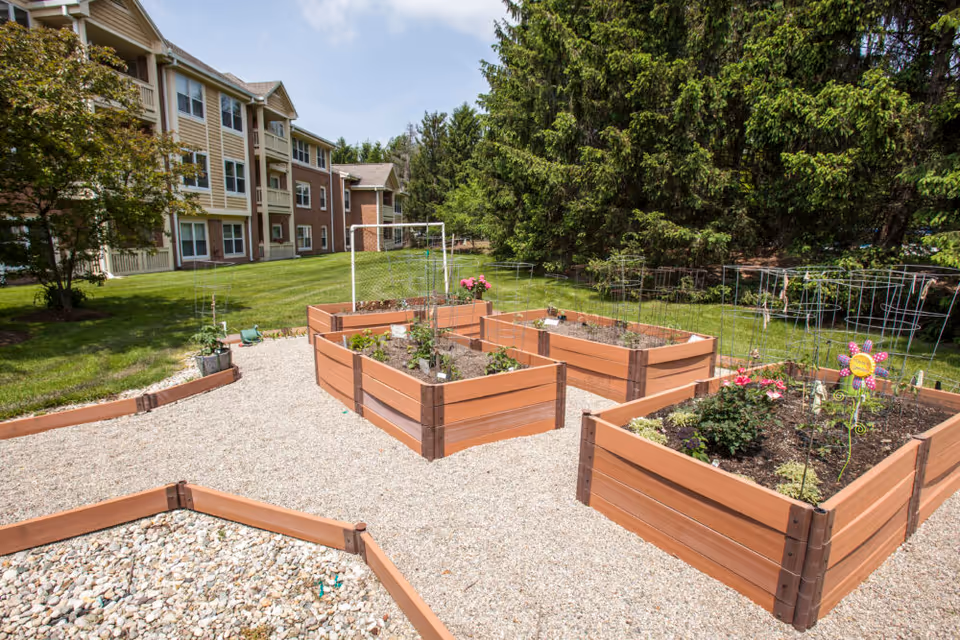 Outdoor garden area with raised wooden garden beds containing various plants and flowers, situated on a gravel surface. In the background, there is a multi-story residential building with beige and brick exterior walls, surrounded by green grass and tall trees under a partly cloudy sky.