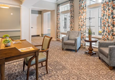 A cozy interior room with patterned carpet, two gray upholstered armchairs, a small round wooden table with a potted plant, a wooden desk with a chair, and large windows with floral curtains allowing natural light to enter.
