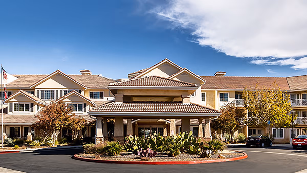 Front exterior of a multi-story senior living facility with a porte-cochère entrance, landscaped roundabout, and parked cars under a blue sky.