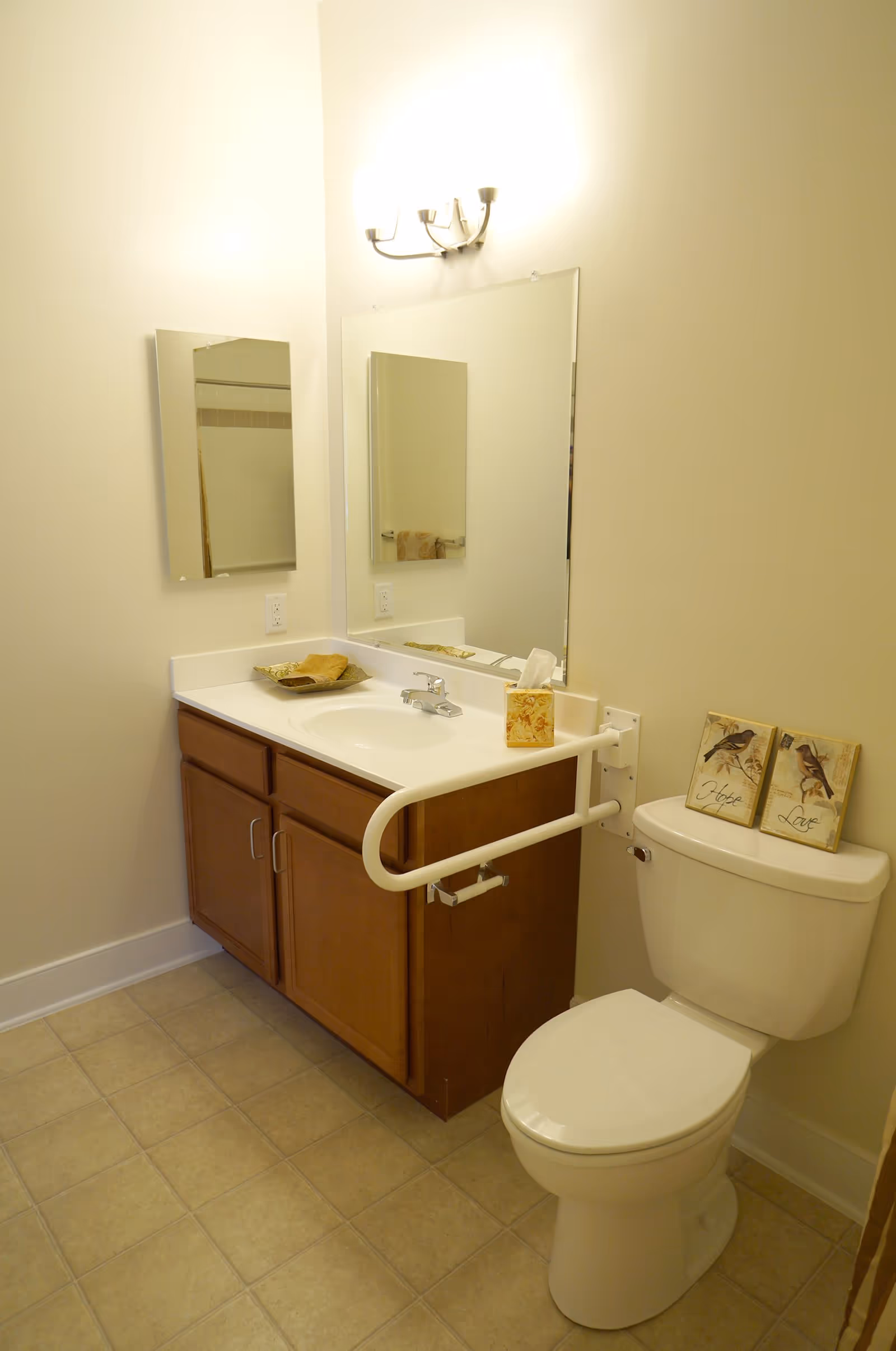 A clean bathroom with a white toilet and a wooden vanity with a white countertop and sink. Above the sink is a large mirror and a smaller medicine cabinet mirror on the adjacent wall. A white safety grab bar is mounted on the side of the vanity next to the toilet. Two decorative plaques with bird images and the words 'Hope' and 'Love' are placed on top of the toilet tank. The floor is tiled in a light beige color, and the walls are painted a light cream color.