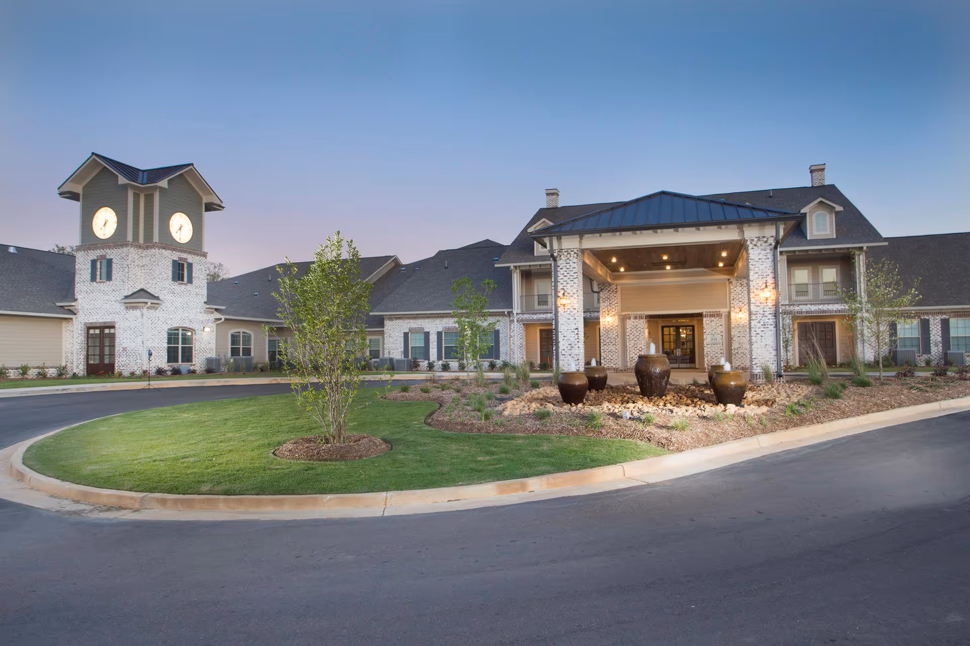 Front entrance of a senior living facility featuring a clock tower, covered porte-cochère, landscaped roundabout and decorative urns.