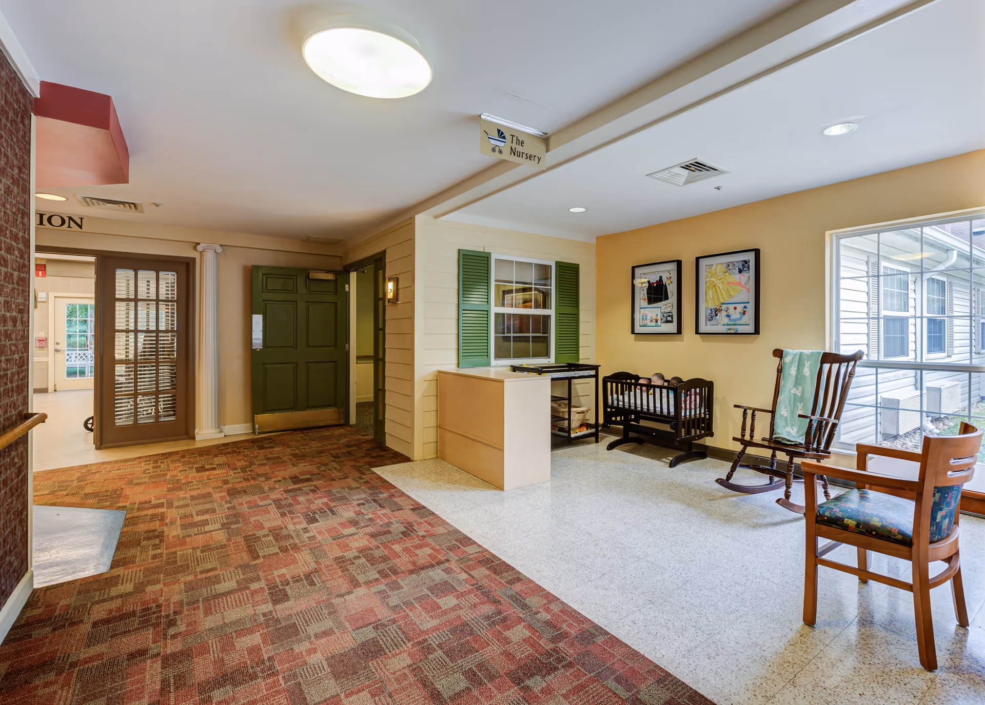 Interior hallway area of a senior living facility with a green door, a small reception desk, a rocking chair, a wooden chair, a baby crib, and framed pictures on the wall. There is a sign above the desk that reads 'The Nursery'. The floor is a combination of patterned carpet and light-colored tile, and there is a large window letting in natural light.