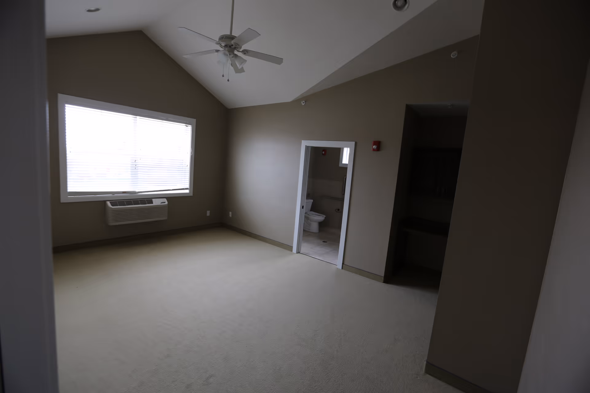 Empty beige room with a vaulted ceiling and ceiling fan, a large window with blinds, wall air conditioner, and an open doorway showing a bathroom with a toilet.
