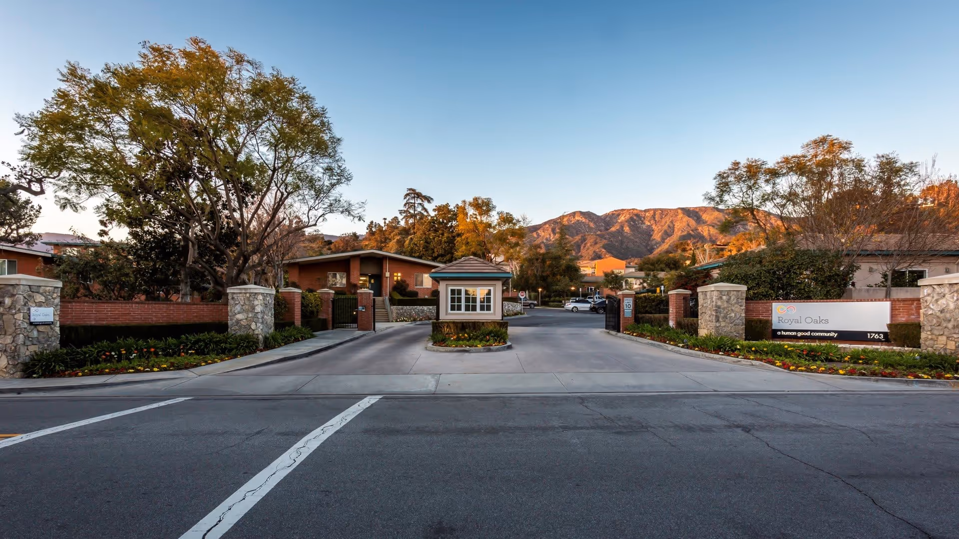 Entrance to the Royal Oaks senior living facility with a gated driveway, a small guardhouse in the center, stone pillars, landscaped flower beds, and mountains in the background under a clear sky.