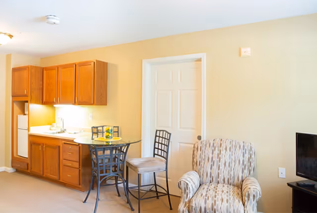 Small senior living studio interior with a kitchenette, round glass dining table with chairs, upholstered armchair, and a TV.