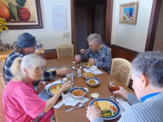 Four elderly people sitting around a wooden dining table eating a meal together in a cozy dining room with framed artwork on the walls.