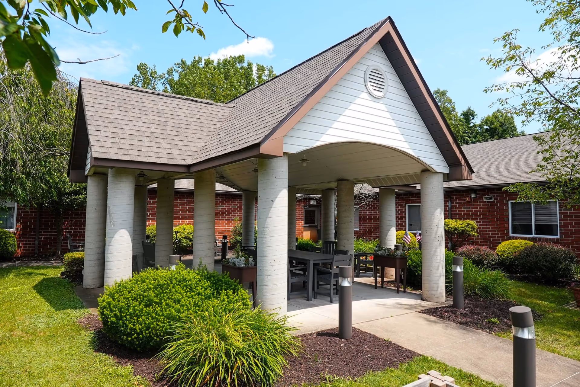 Outdoor covered seating area with a peaked roof supported by large cylindrical columns, surrounded by green bushes and trees, with a brick building in the background under a clear blue sky.