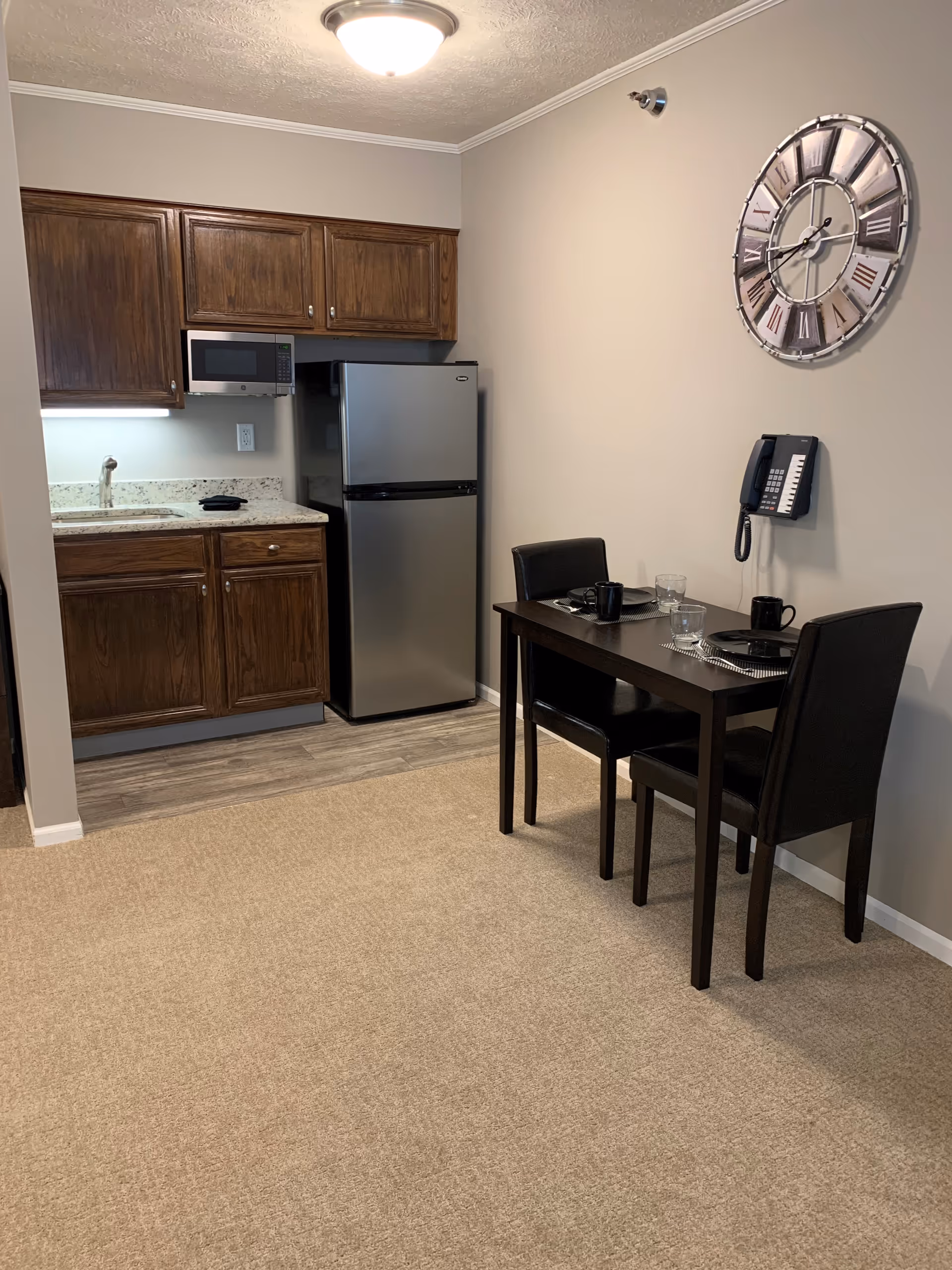 Small kitchen area with wooden cabinets, a stainless steel refrigerator, and a microwave above the counter. Adjacent to the kitchen is a small dining table set for two with black chairs, black mugs, and glasses. A large decorative wall clock and a wall-mounted telephone are visible on the beige wall.