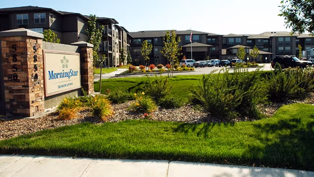 Exterior view of MorningStar Senior Living of Boise facility showing a large, multi-story building with multiple windows, a well-maintained green lawn, shrubs, and a stone sign with the facility's name. Several cars are parked in front of the building under a clear sky.