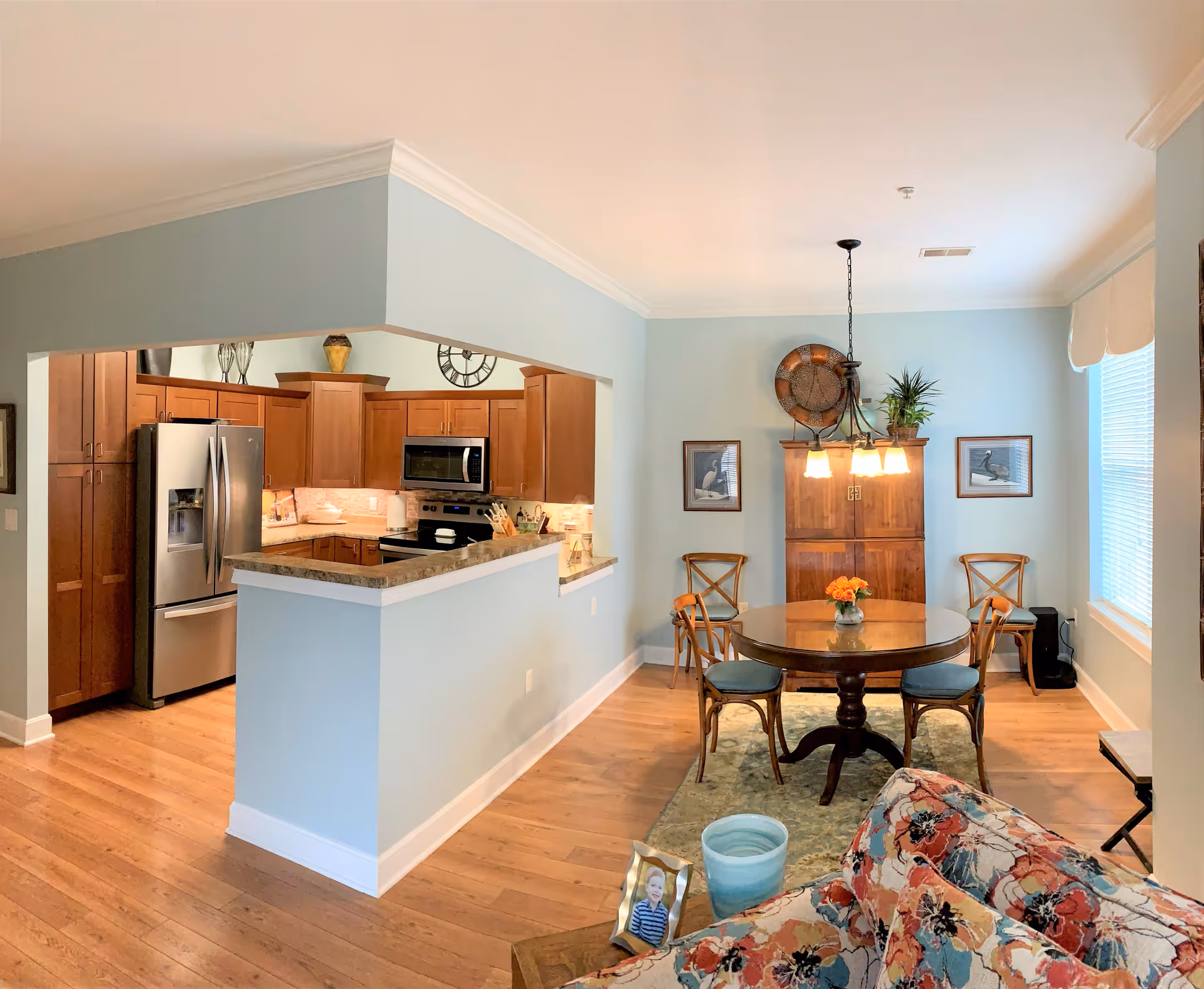 A bright and cozy interior space featuring a kitchen and dining area. The kitchen has wooden cabinets, stainless steel refrigerator, microwave, and stove. The dining area includes a round wooden table with four chairs, a hanging light fixture, and a wooden cabinet against the wall with decorative items and framed pictures. The floor is wooden, and there is a floral patterned couch partially visible in the foreground.