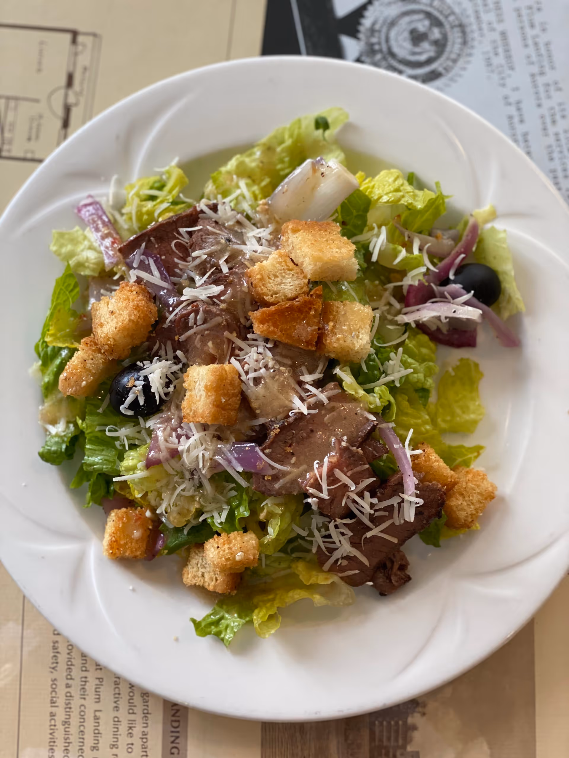 A white plate with a fresh salad consisting of leafy greens, slices of cooked meat, shredded cheese, croutons, black olives, and red onion slices.