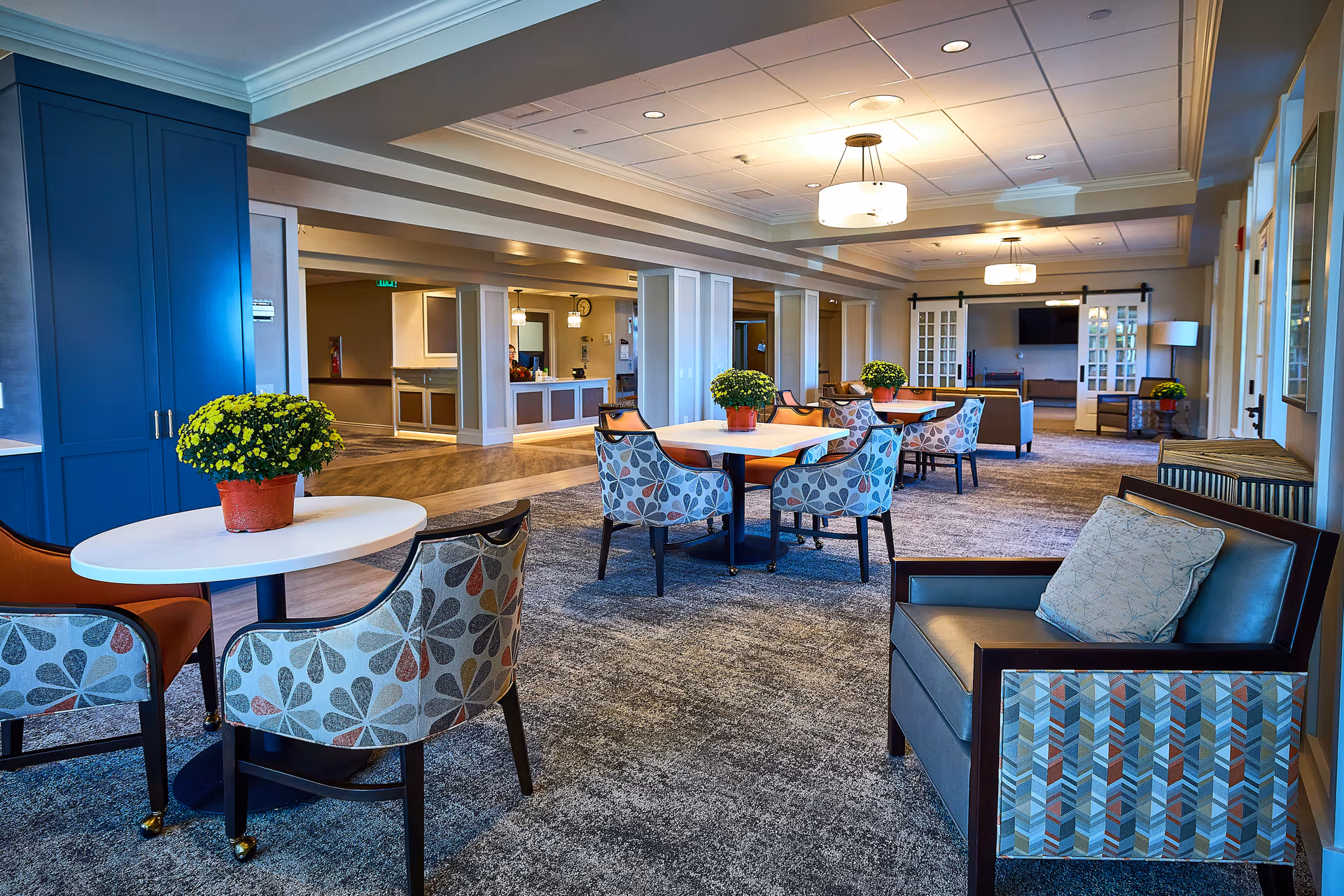 A spacious and well-lit common area in a senior living facility featuring multiple tables with patterned chairs and potted yellow flowers on each table. The room has carpeted flooring, modern ceiling lights, and a seating area with armchairs and a TV in the background. There is a reception desk visible towards the back left side of the room.
