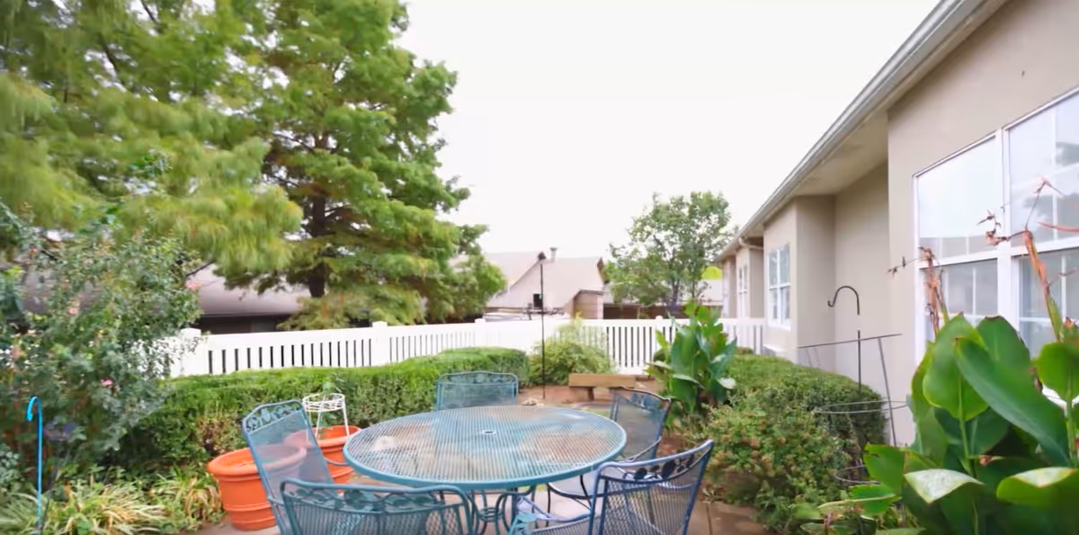 Outdoor patio area with a round metal table and six matching chairs surrounded by green plants and bushes. A white picket fence encloses the space, and there are large trees and a beige building with windows in the background.