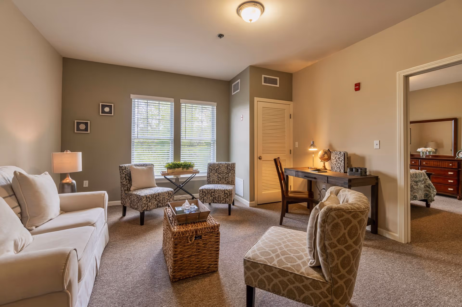 A cozy living room area with a white sofa, two patterned armchairs, a wicker coffee table, and a small table with a plant in front of two windows with blinds. There is a wooden desk with a chair, a globe, and a lamp against the wall. The room has beige walls and carpeted floor. An open doorway leads to a bedroom with a dresser and mirror.
