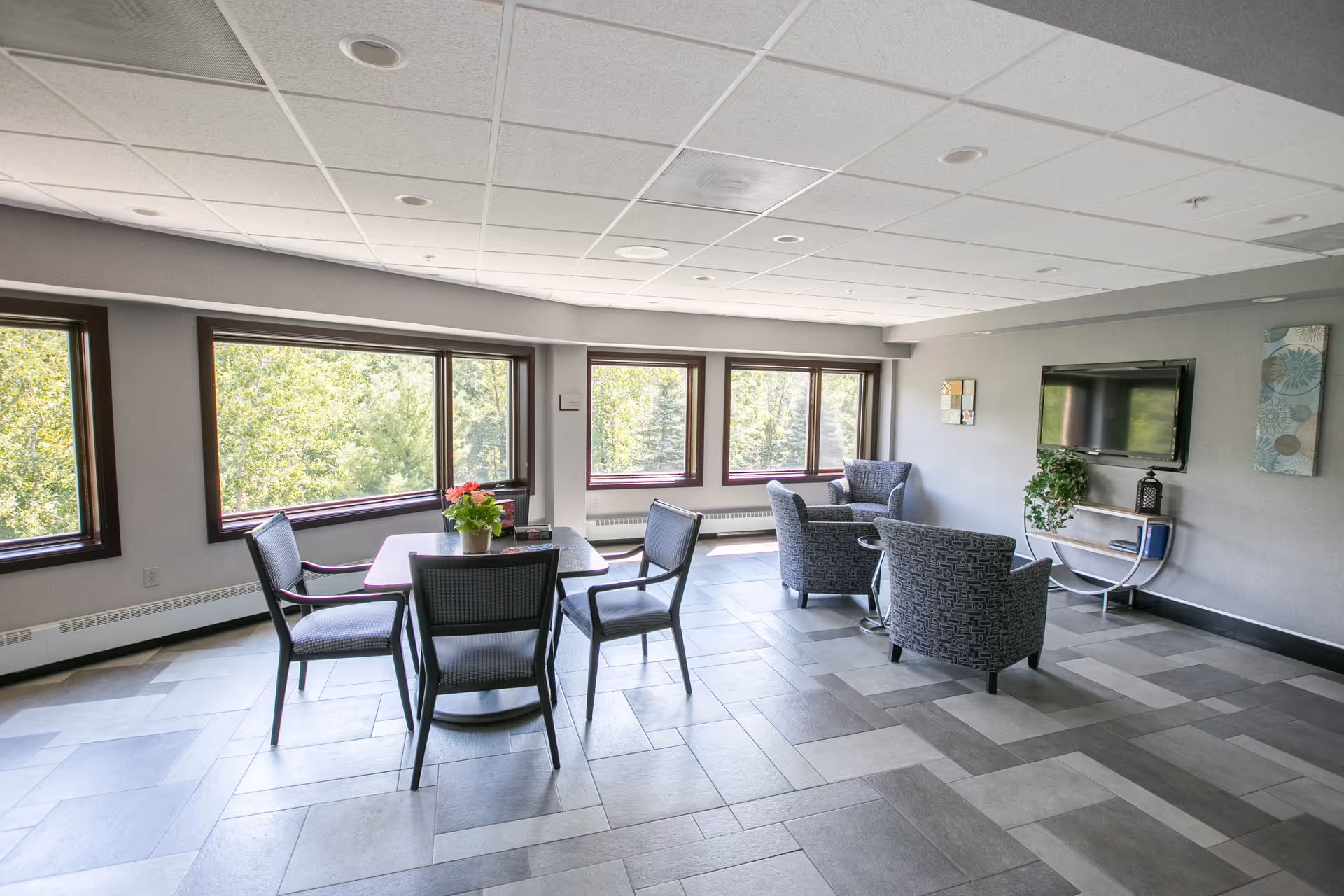 A bright and spacious common area with large windows showing green trees outside. The room has a tiled floor and a ceiling with recessed lighting. There is a square table with four chairs around it, a small plant on the table, and a seating area with three upholstered armchairs facing a wall-mounted flat-screen TV. The walls are decorated with a few pieces of artwork and a small shelf with a plant and a lantern.