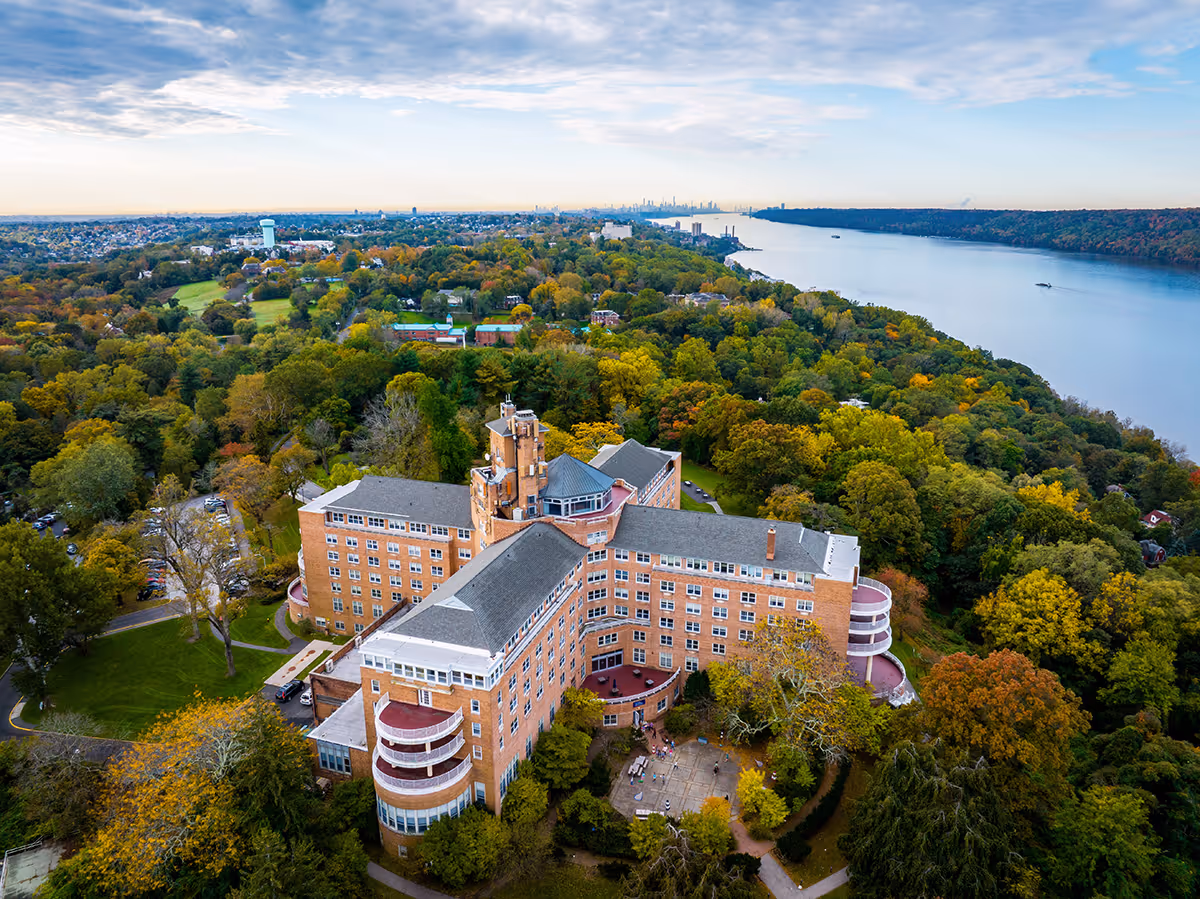 Aerial view of a large multi-story brick building on wooded grounds beside a river.