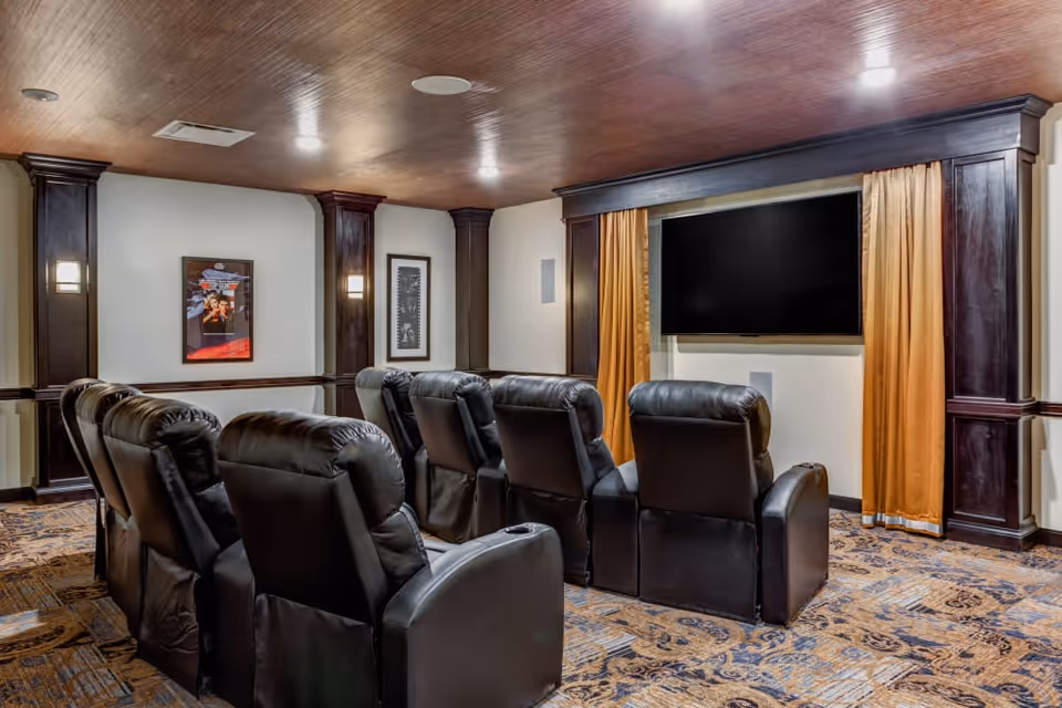 A cozy home theater room with two rows of black leather recliner chairs facing a large flat-screen TV mounted on the wall. The room features dark wood paneling, warm lighting, framed artwork on the walls, and patterned carpet flooring.