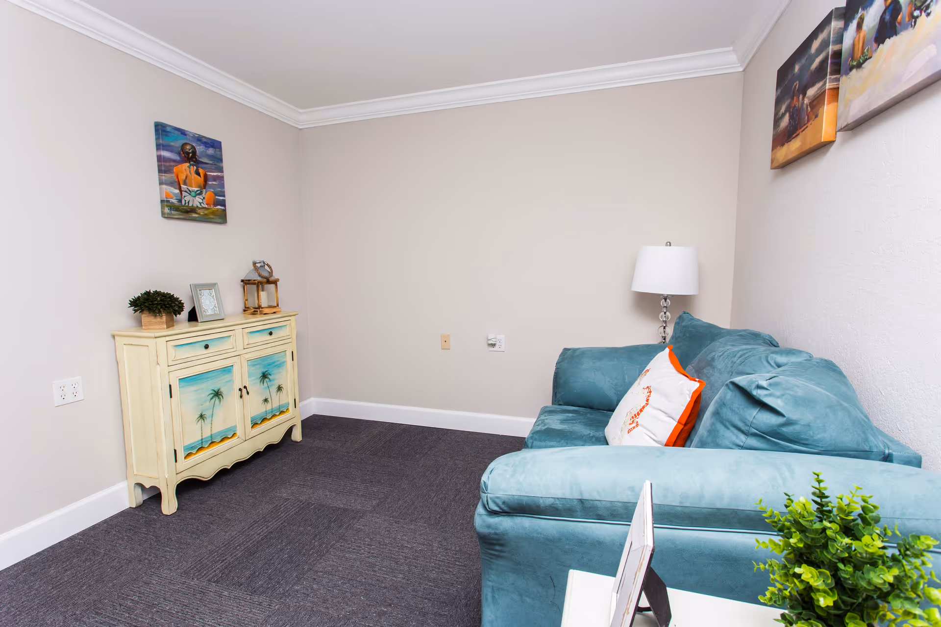 A small living room with a teal couch adorned with a white pillow featuring an orange seahorse design. A white side table with a green plant and a picture frame is in front of the couch. On the opposite wall, there is a cream-colored cabinet with painted palm tree designs and decorative items on top. The walls are light beige with three colorful paintings hanging above the couch and cabinet. The floor is covered with dark gray carpet tiles.