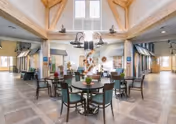Spacious and bright common area in a senior living facility with high vaulted ceilings and exposed wooden beams. Several round tables with chairs are arranged in the center, decorated with small plants and flowers. Large windows allow natural light to fill the room, and there are multiple seating areas visible in the background.