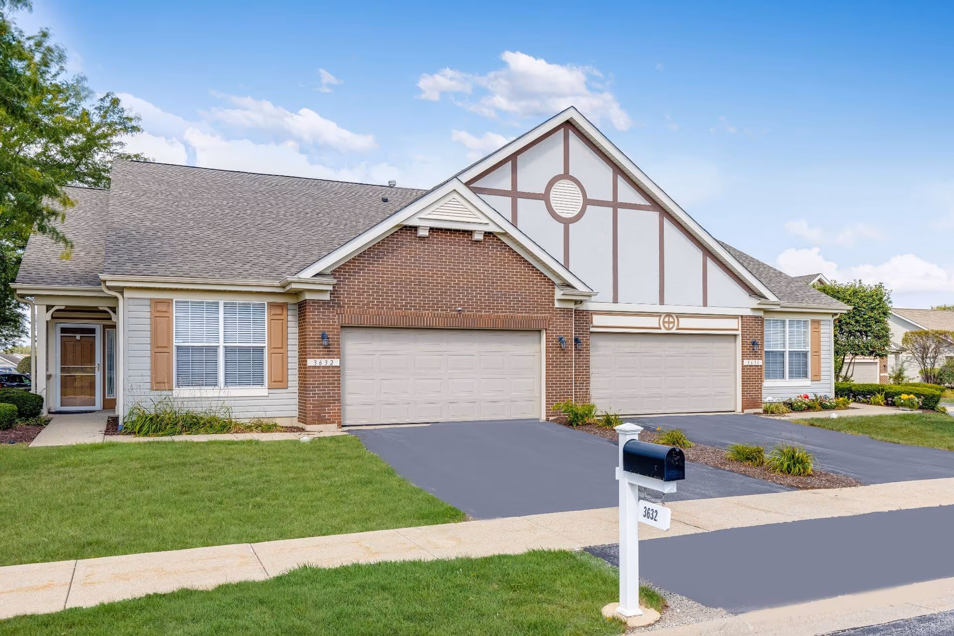 Exterior view of a residential-style building with two attached garages, brick and siding facade, and a mailbox labeled 3632 in front. The building has a well-maintained lawn and a clear blue sky with some clouds in the background.