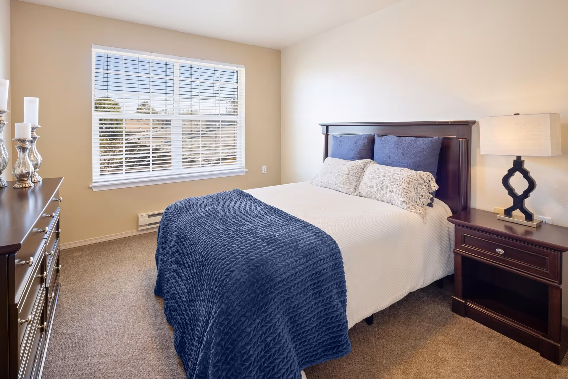 A neatly made bed with a dark wooden headboard, white bedding, blue pillows, and a blue textured blanket in a bright bedroom with beige walls and carpet. There is a wooden nightstand with a lamp on the right side of the bed and a wooden dresser with decorative candle holders on the left side. A large window with white blinds lets in natural light.
