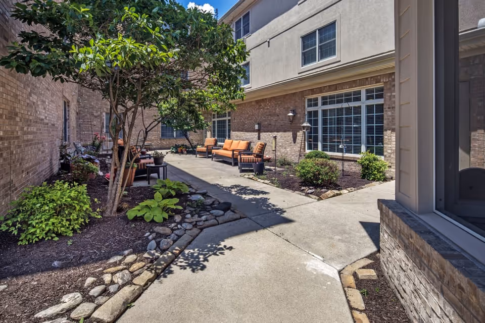 Sunlit enclosed courtyard with patio seating, landscaped plant beds, and a concrete walkway alongside a brick building.