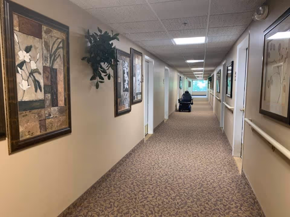 Long carpeted hallway with framed artwork and handrails, ending with a person in a mobility scooter.