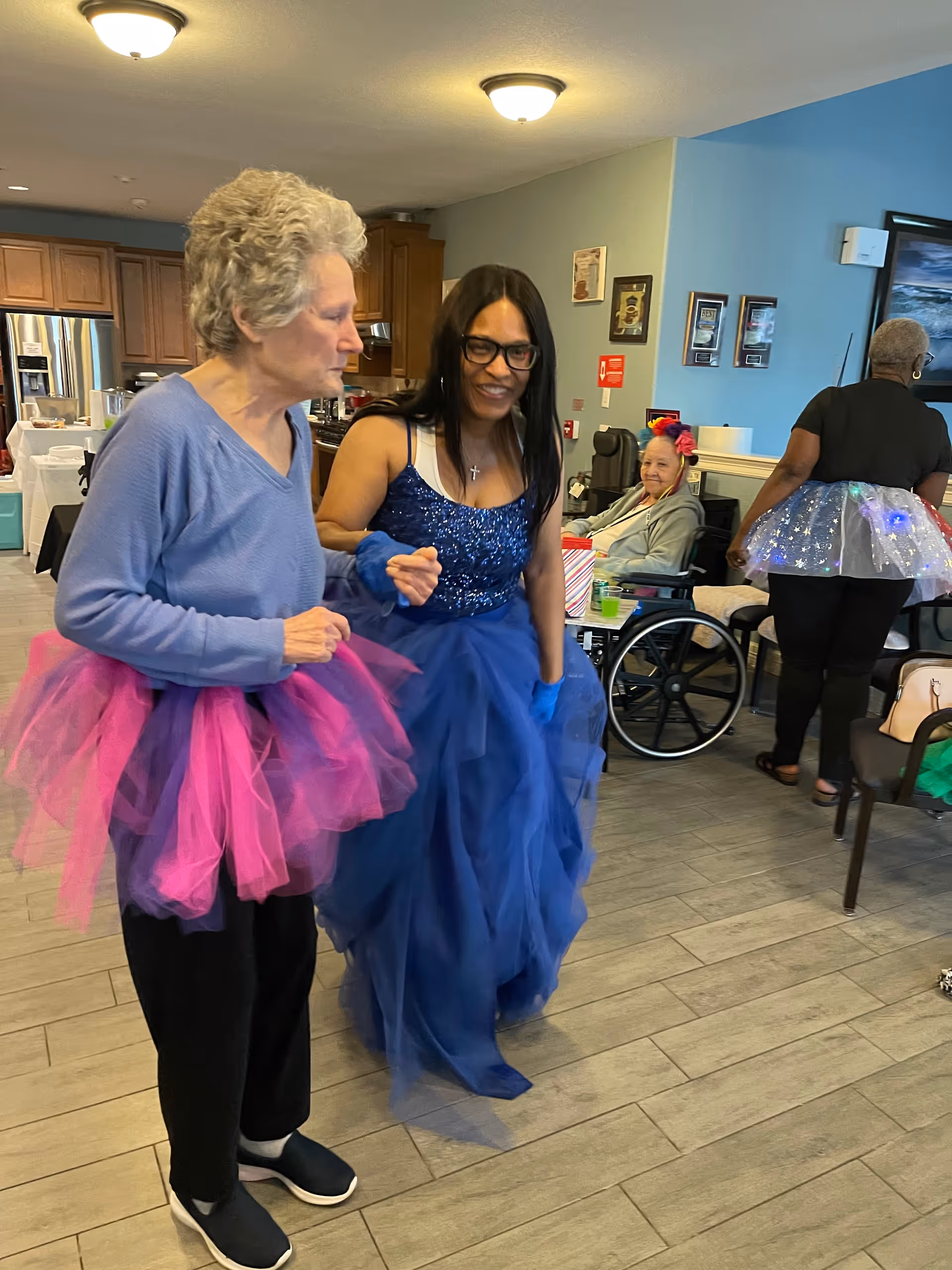 Two women wearing colorful tutus and a blue gown dance together in a communal room with other people, wheelchairs, and a kitchen area in the background.