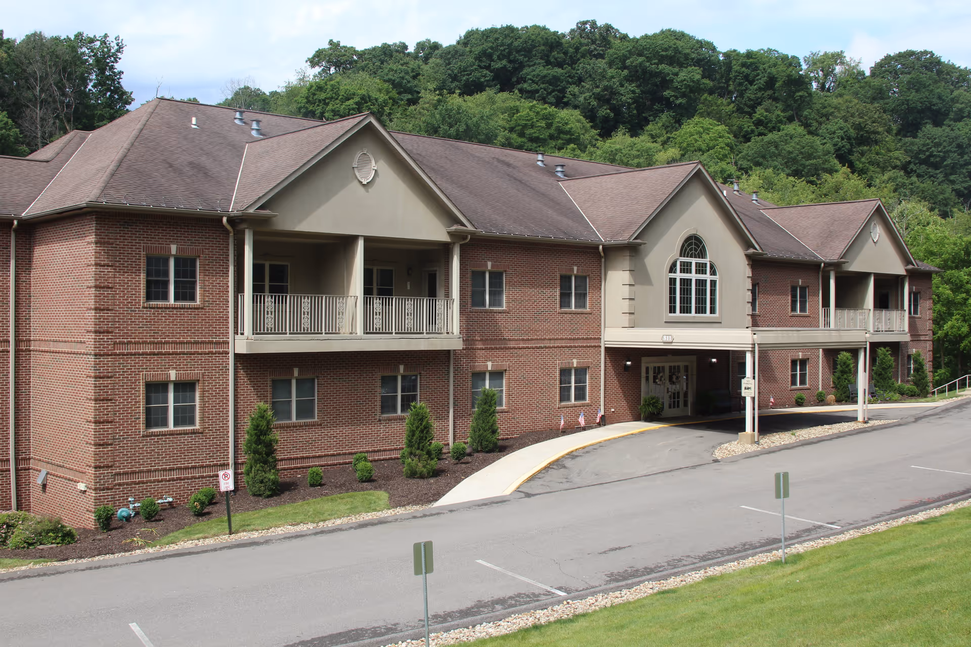 Exterior view of a two-story brick building with beige accents, balconies, and a covered entrance, surrounded by greenery and a parking area in front.