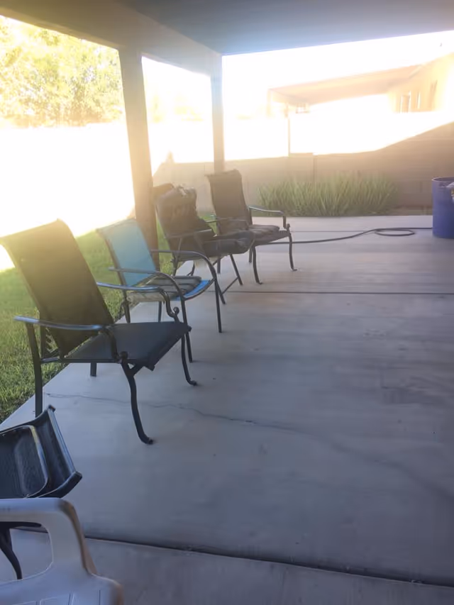 Covered outdoor patio area with several metal and plastic chairs arranged in a row on a concrete floor. There is a grassy area and a block wall in the background with some plants and a blue trash bin visible.