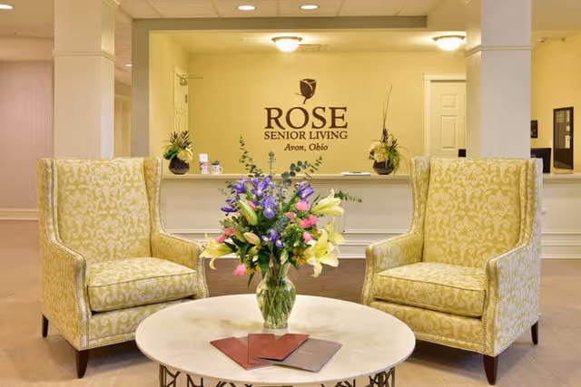 Lobby reception area with two patterned armchairs flanking a round table with a vase of flowers and a reception desk bearing the 'Rose Senior Living Avon, Ohio' logo on the wall.