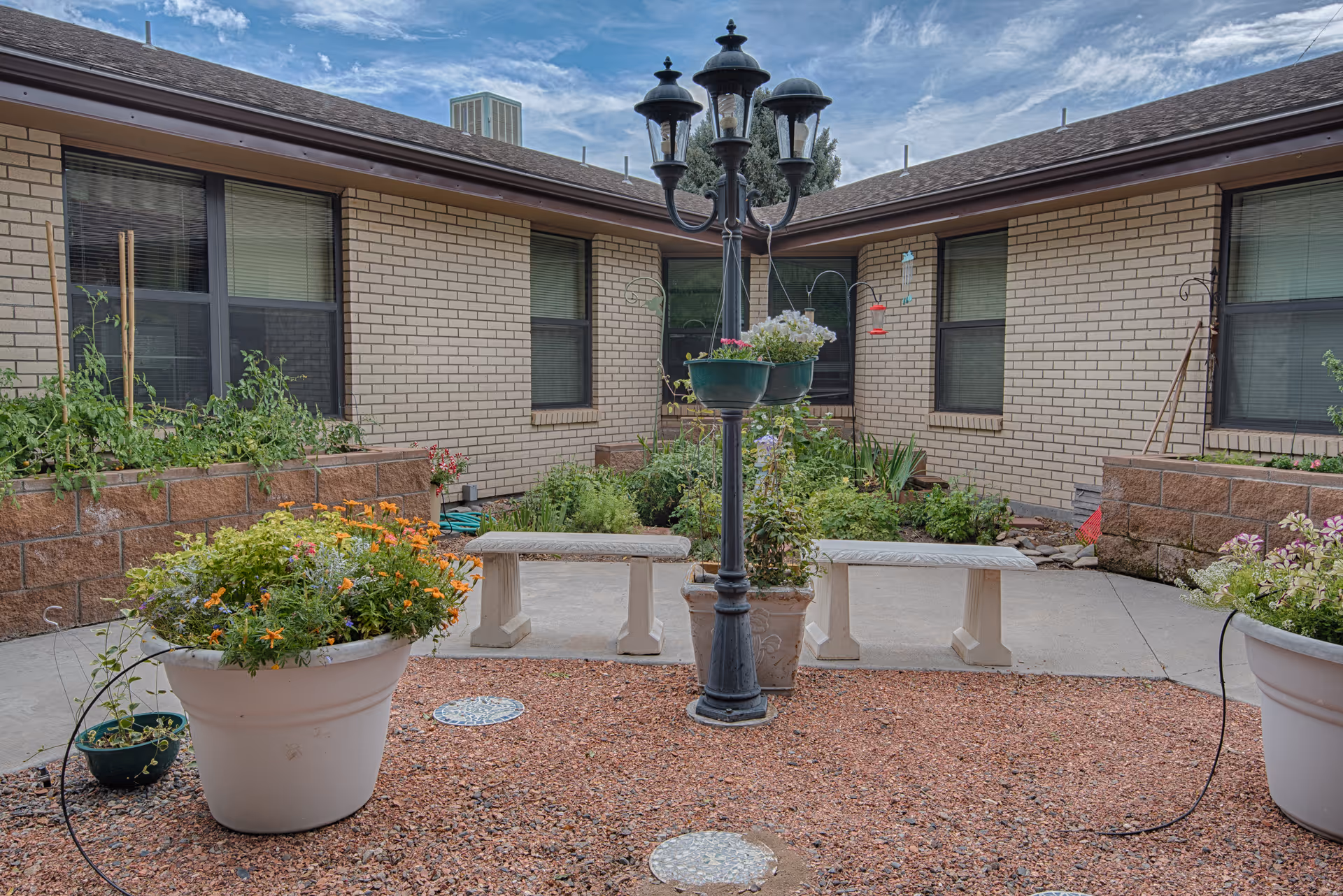 Outdoor courtyard area with a central black lamppost adorned with hanging flower pots, surrounded by two white stone benches. The courtyard is bordered by raised garden beds with various plants and flowers, set against a beige brick building with several windows. The ground is covered with reddish gravel and concrete walkways under a partly cloudy sky.