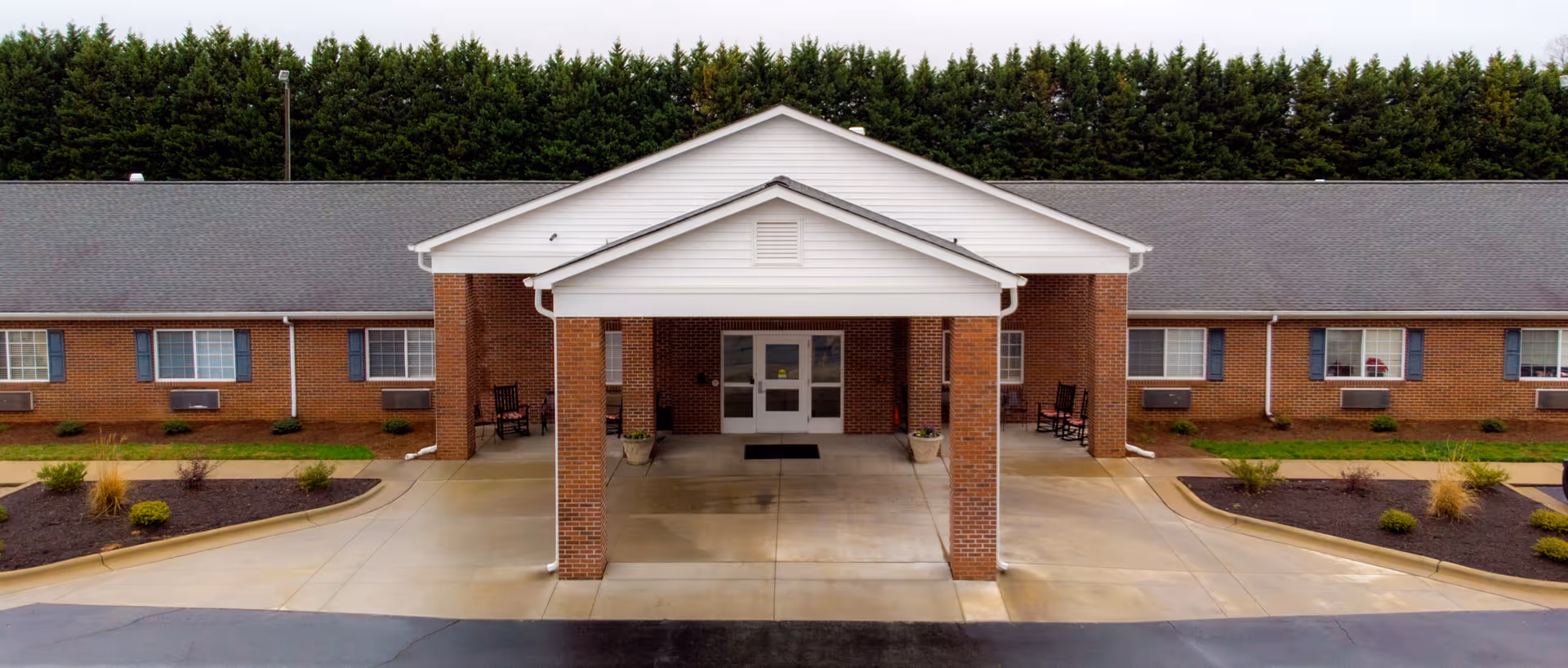 Front entrance of a single-story brick senior living building with a covered portico, driveway, and landscaped beds.