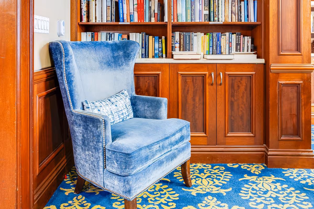 A cozy blue velvet armchair with a patterned cushion is placed in front of a wooden bookshelf filled with books. The room features wood paneling on the walls and a blue carpet with yellow floral patterns.