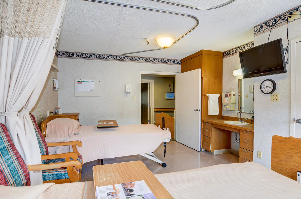 Senior care patient room with two beds, bedside table, wall-mounted TV, sink and cabinetry.