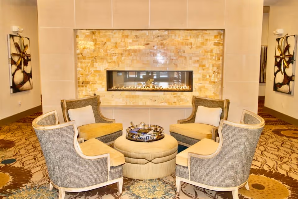 A cozy seating area with four cushioned armchairs arranged around a round ottoman in front of a modern rectangular gas fireplace set in a wall with beige and cream stone tiles. The room features patterned carpet and two abstract floral paintings on the walls.