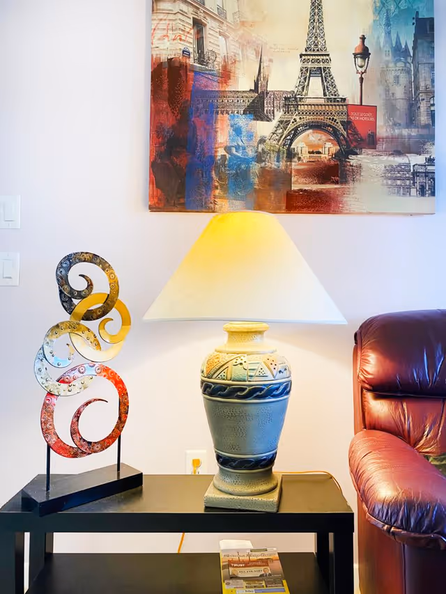 A cozy corner featuring a black side table with a decorative metal sculpture and a ceramic table lamp with a beige shade. Above the table hangs a colorful artwork depicting the Eiffel Tower and other Parisian elements. To the right, part of a brown leather armchair is visible.