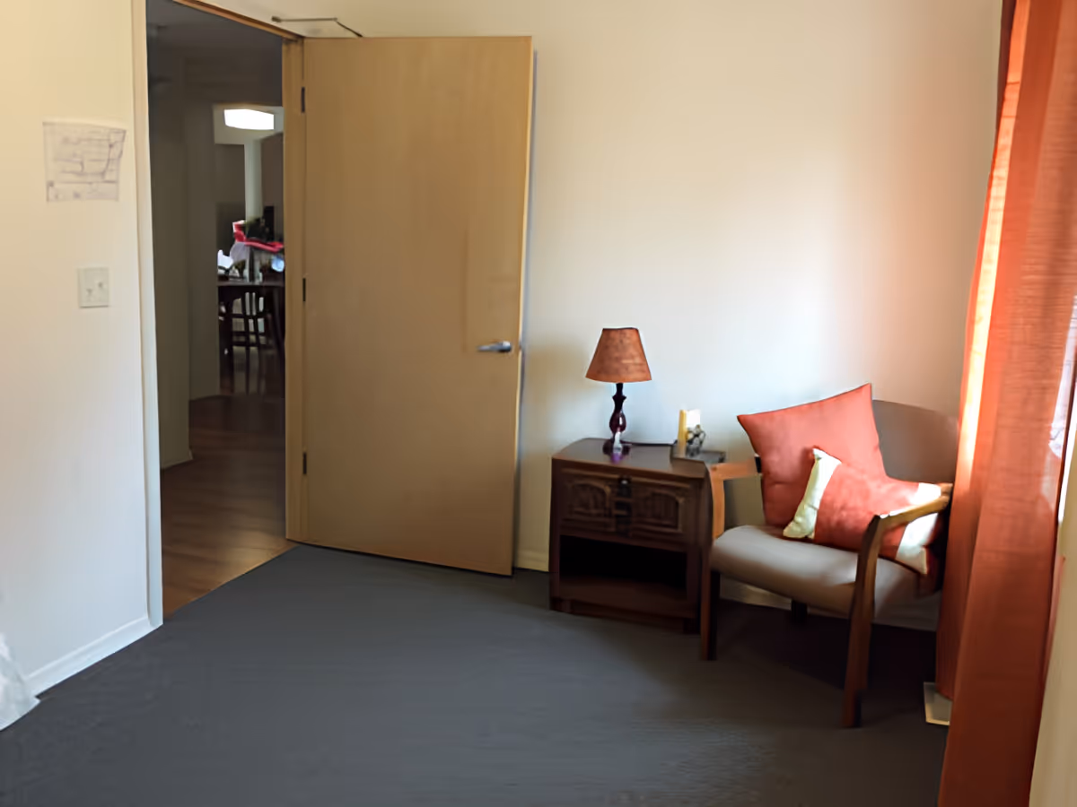 Small sitting area with an upholstered chair, side table and lamp beside an open wooden door and orange curtains.