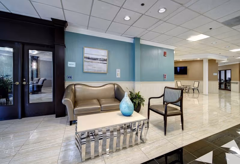 A seating area in a senior living facility with a brown leather loveseat, a wooden chair with striped upholstery, and a modern coffee table with a blue decorative vase. The walls are painted blue and beige, with a framed abstract painting hanging above the loveseat. There are double glass doors to the left and a hallway with additional seating and a wall-mounted TV in the background.