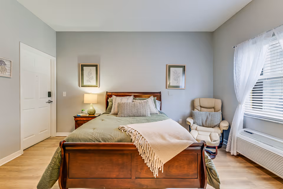 Well-lit bedroom with a wooden bed made up in green bedding, a nightstand with a lamp, a recliner by a curtained window, and light wood floors.