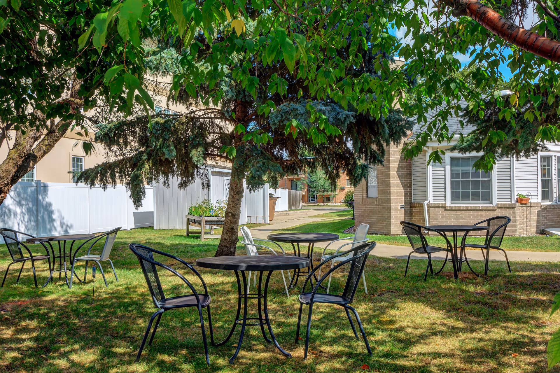 Outdoor courtyard with metal tables and chairs under leafy trees at a senior living facility.