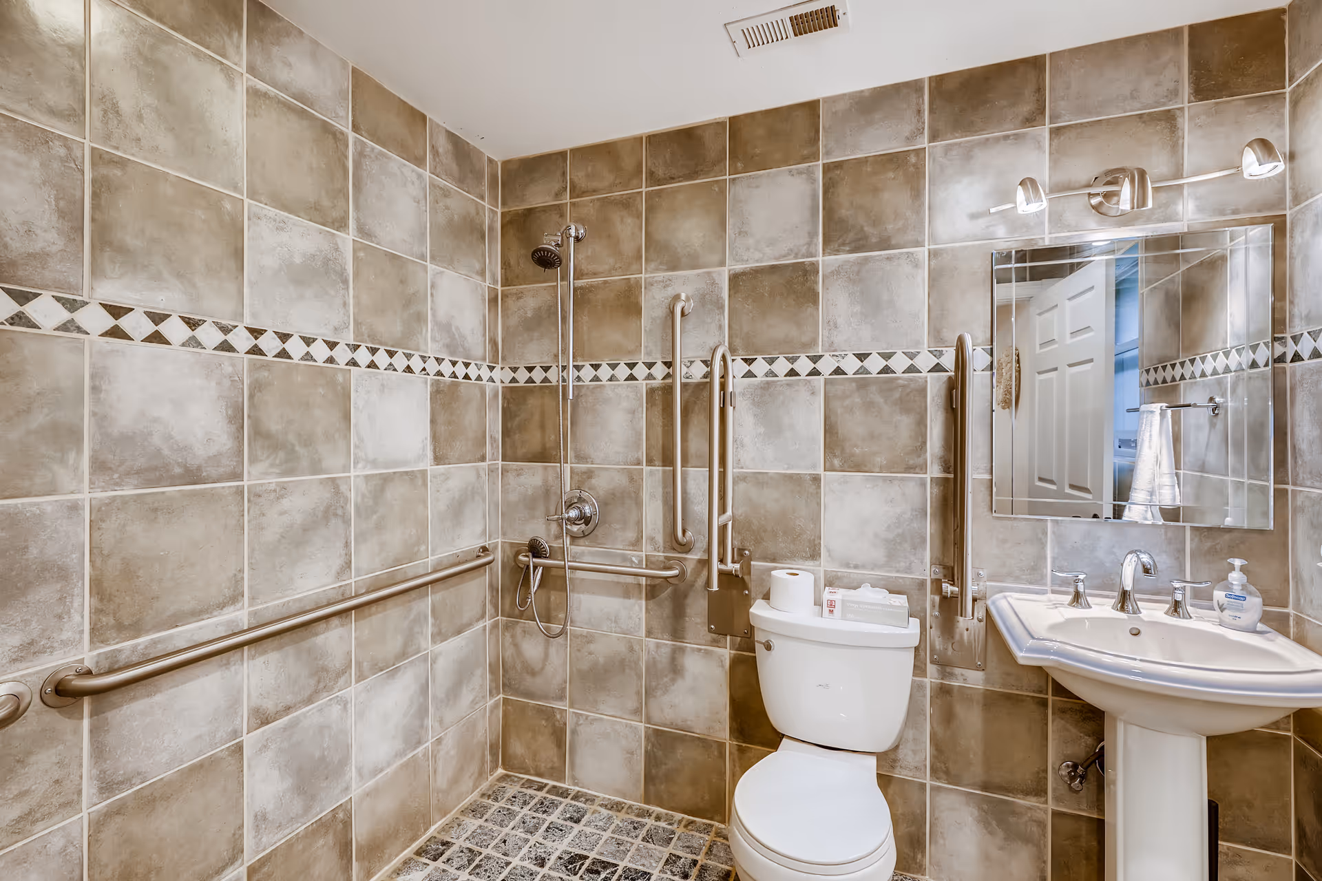 Accessible bathroom with tiled walls featuring a walk-in shower, grab bars, toilet, and pedestal sink with mirror.