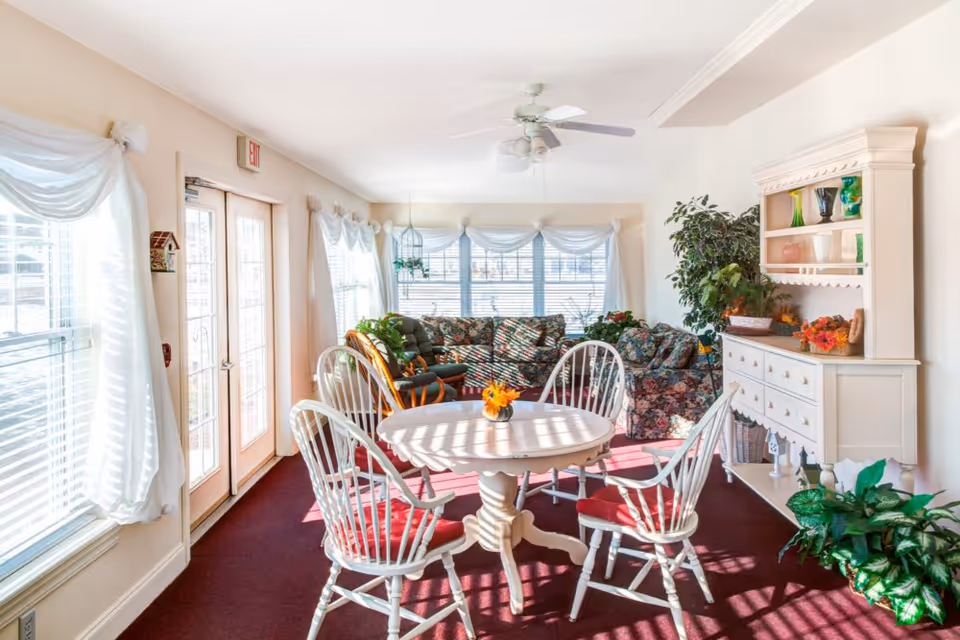 A bright and cozy living room area with a round white table surrounded by four white chairs with red cushions. Behind the table, there are floral-patterned couches and a white hutch with decorative items. Large windows with white curtains allow natural light to fill the room, and there are several green plants adding a touch of nature.