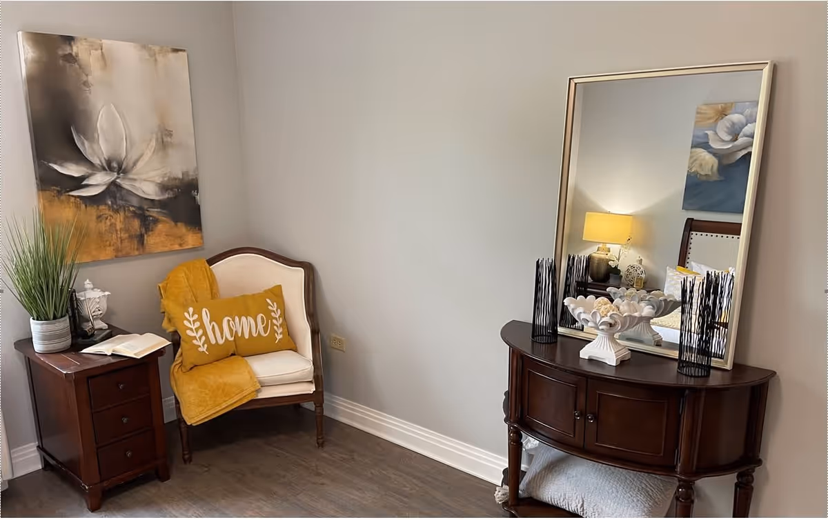 A cozy corner of a room featuring a wooden armchair with a white cushion, a yellow throw blanket, and a yellow pillow with the word 'home' on it. Next to the chair is a small wooden side table with a potted plant and an open book. On the adjacent wall hangs a large painting of a white flower with a yellow and black background. Across from the chair is a dark wooden console table with a large mirror above it, decorative vases, and a bowl filled with decorative spheres. The room has light gray walls and wood flooring.