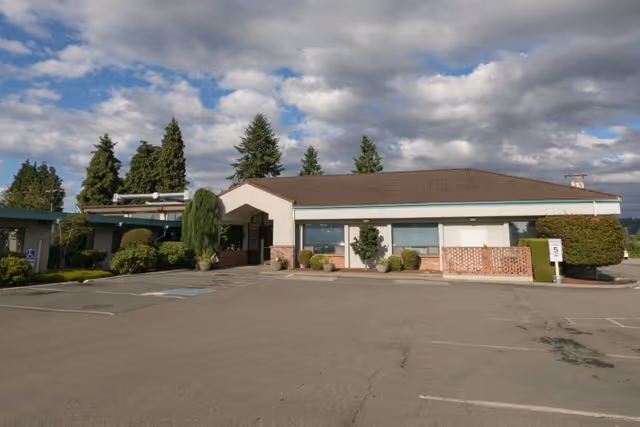 Single-story building with an entrance, empty parking lot, and shrub landscaping under a partly cloudy sky.