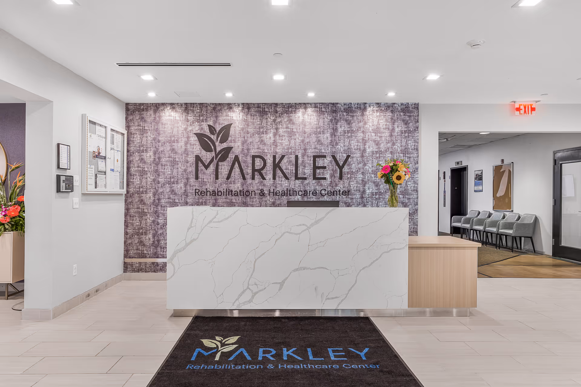 Reception area of Markley Rehabilitation & Healthcare Center featuring a white marble-patterned front desk, a purple textured accent wall with the facility's name and logo, a vase with colorful flowers on the desk, a black floor mat with the facility's name and logo, and a hallway with chairs and an exit sign in the background.