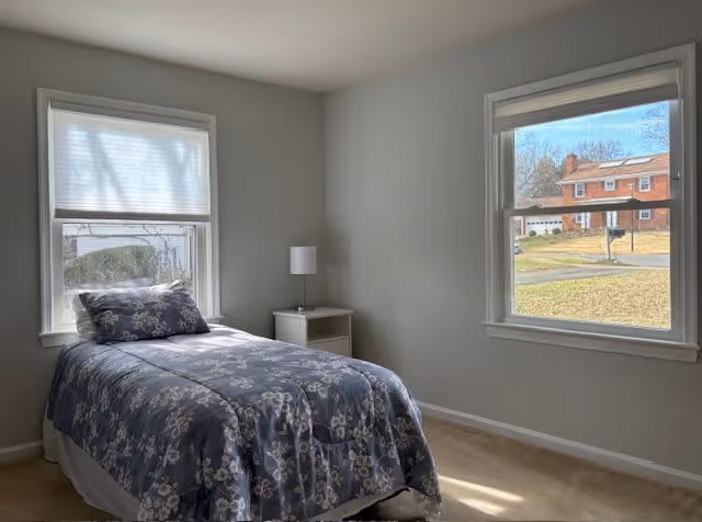 A simple bedroom with a single bed covered in a blue floral comforter and matching pillow. The room has light gray walls, beige carpet, two windows with white blinds, and a small white nightstand with a white lamp between the windows.