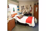 A small, cozy bedroom in a senior living facility with a single bed covered in white sheets and a red blanket. The room has a window with blinds, a wooden cabinet, and various personal decorations and pictures on the walls and surfaces.