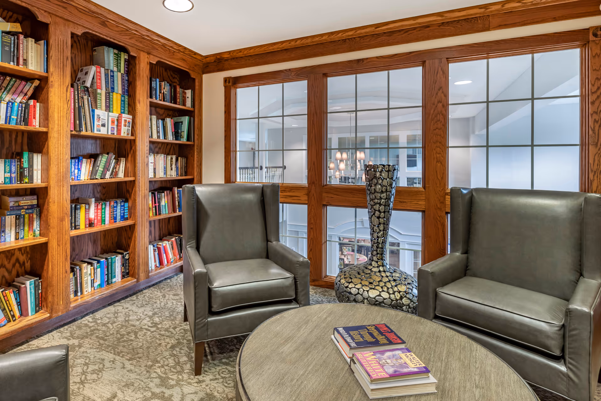 Cozy reading nook in a senior living facility with wooden bookshelves filled with books, two dark leather armchairs, a round wooden table with books on it, and a decorative tall vase. Large windows with wooden frames overlook an interior atrium with lights and railings.