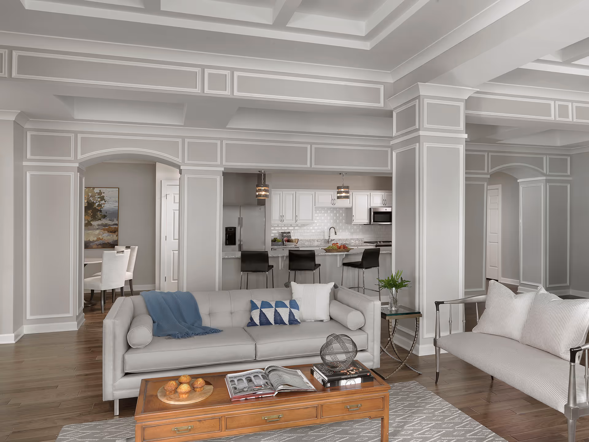 Bright open-plan living room with neutral sofas and a wooden coffee table, looking into a kitchen and dining area with decorative molding.